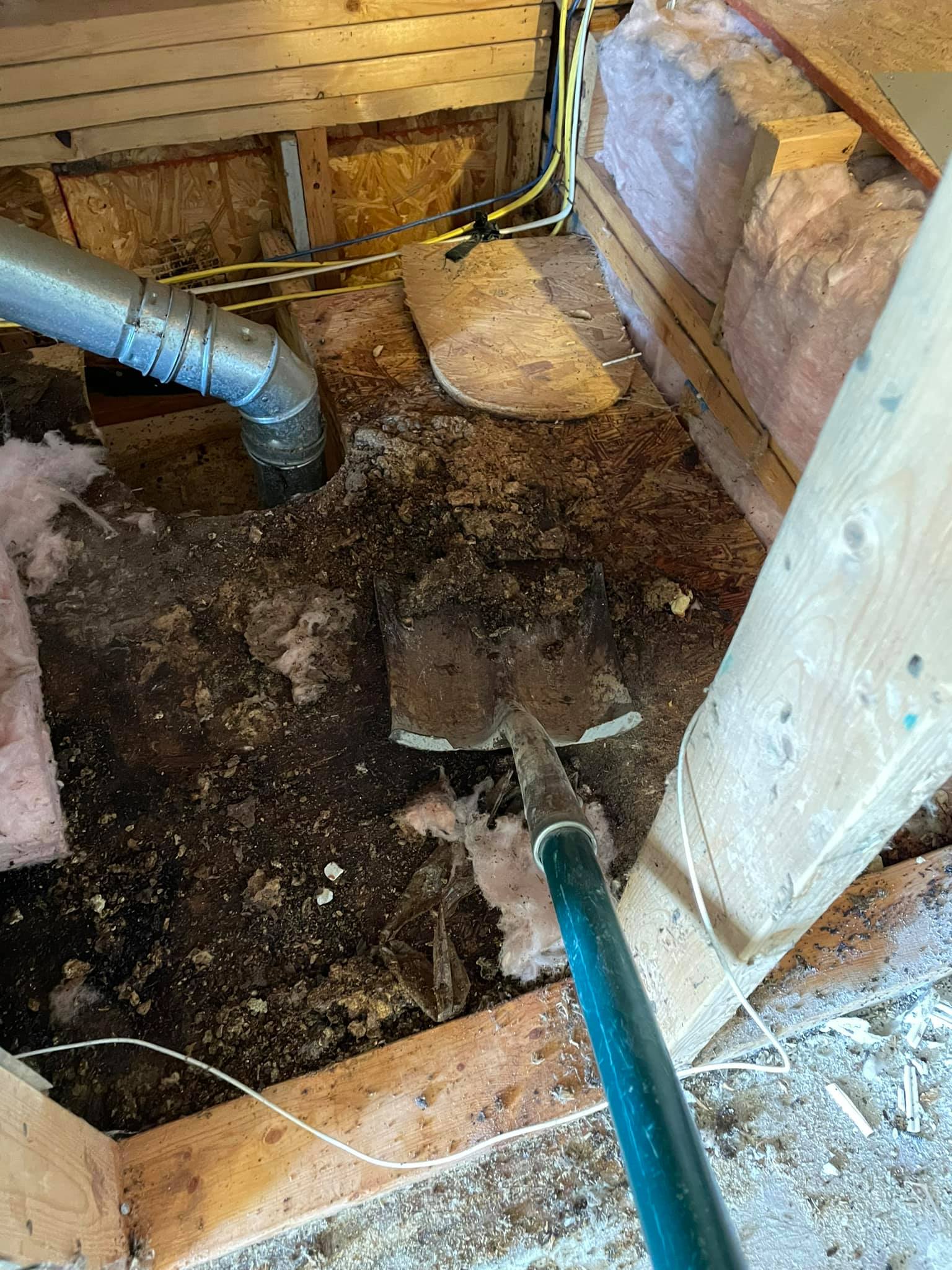 A shovel is positioned on a dirt-covered floor inside an unfinished space with exposed insulation, wooden framing, ducts, and electrical wiring.