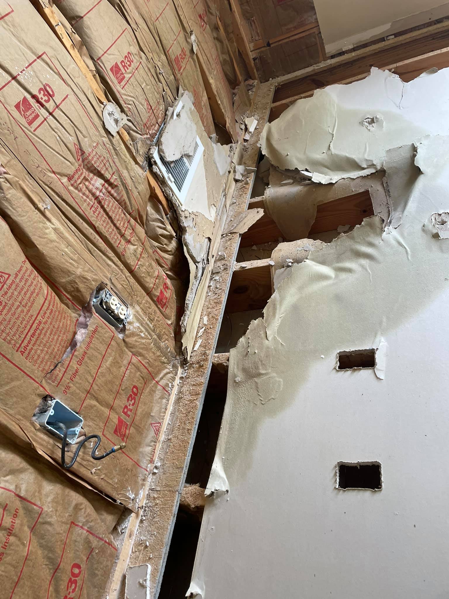 A partially demolished interior wall reveals insulation, exposed wiring, and damaged drywall with holes and broken sections.