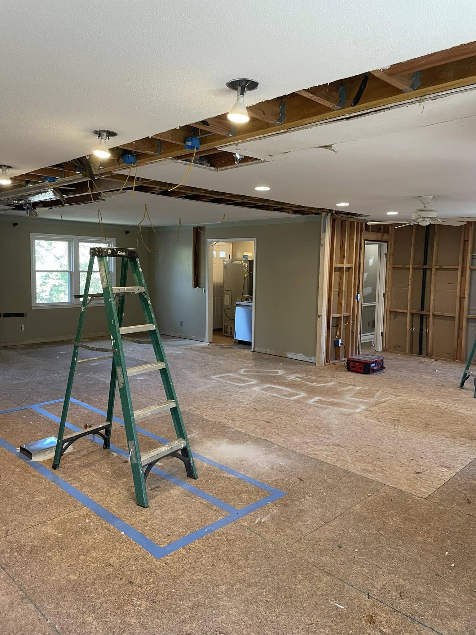 Room under renovation with exposed ceiling beams, partial wall studs, construction materials, and a green ladder in the center. Blue tape marks areas on the floor.