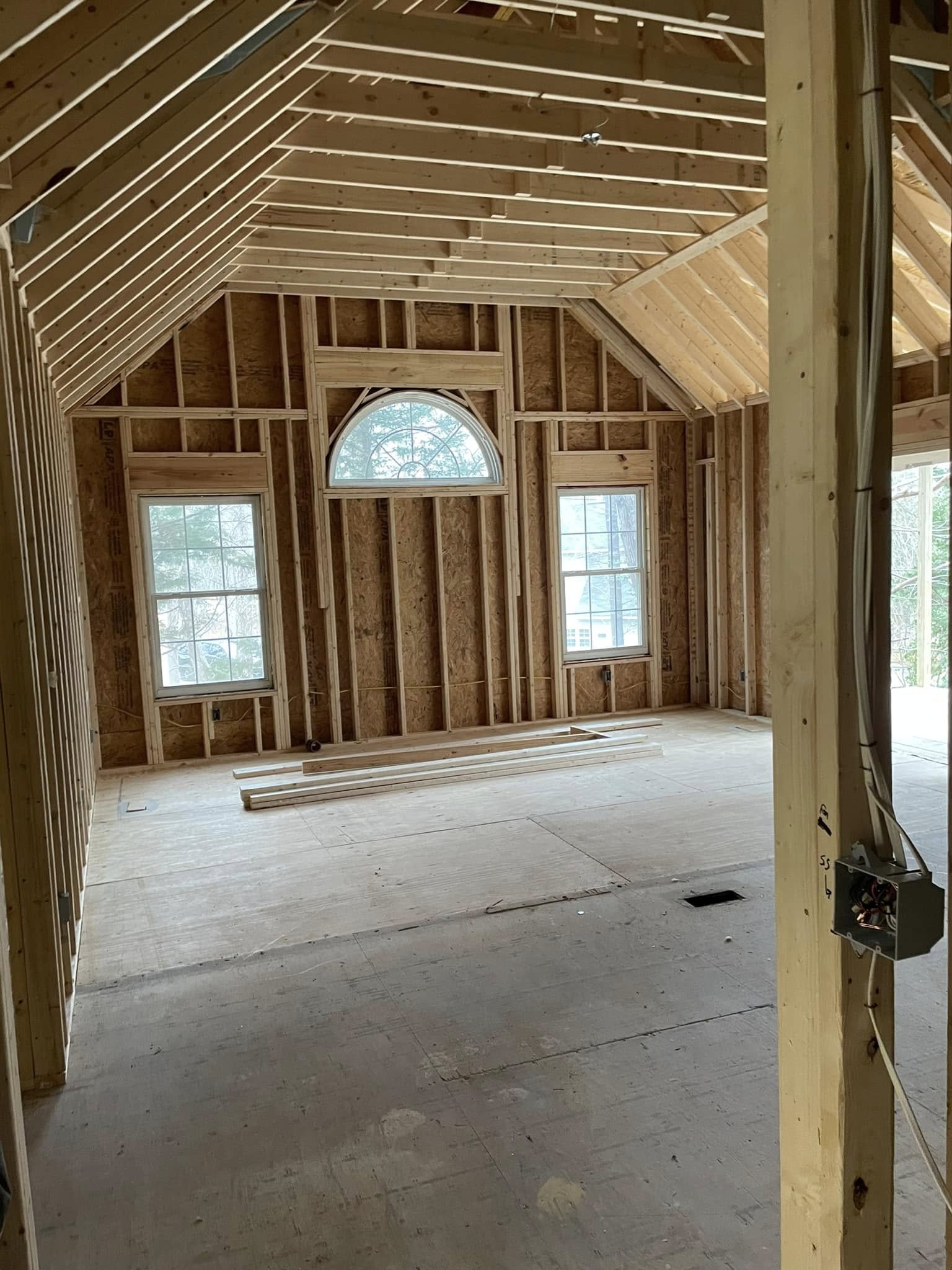 Unfinished room with exposed wooden framing, three windows including an arched center window, and electrical wiring visible on the right.