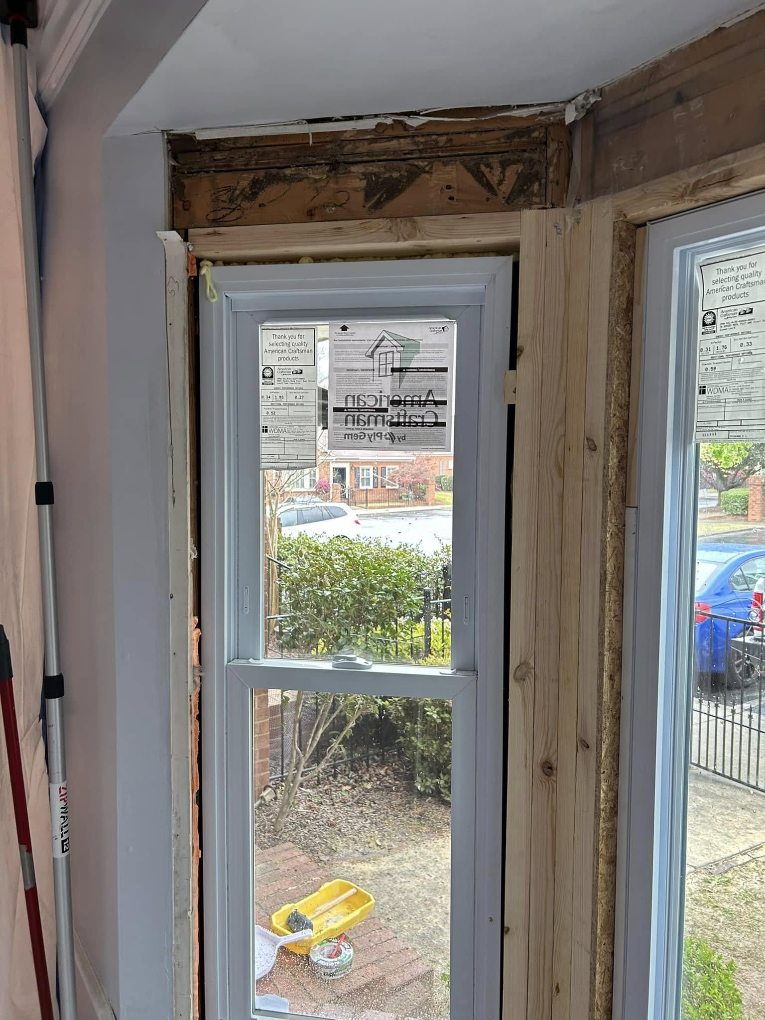 Partially installed window with exposed framing and construction tools visible; view of a street and parked cars outside.