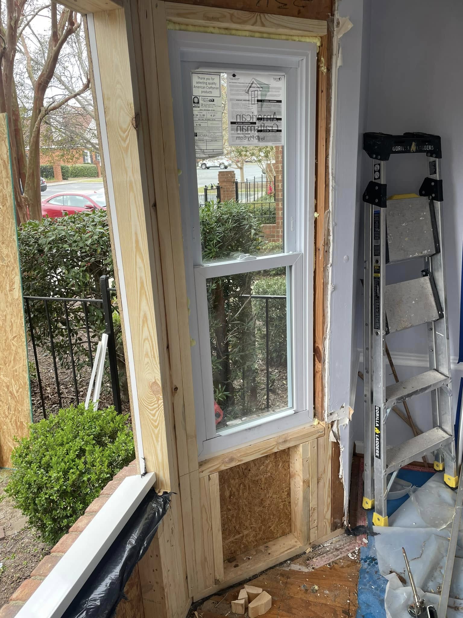 A newly installed window is framed with unfinished wood in a partially renovated wall. A step ladder and renovation materials are visible inside the room.