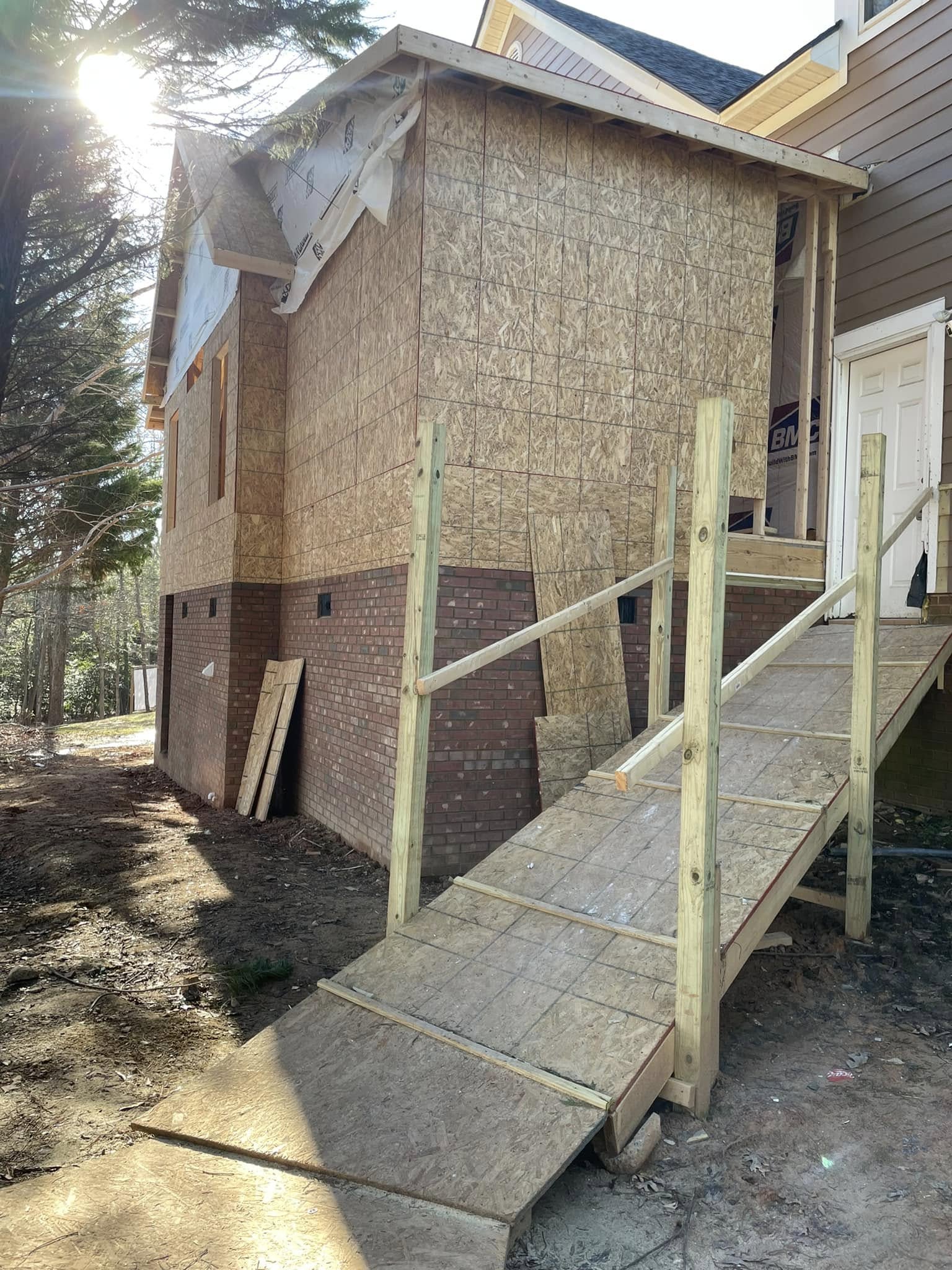 A house under construction features a plywood ramp leading to the door, with exposed plywood walls and brickwork on the lower half.