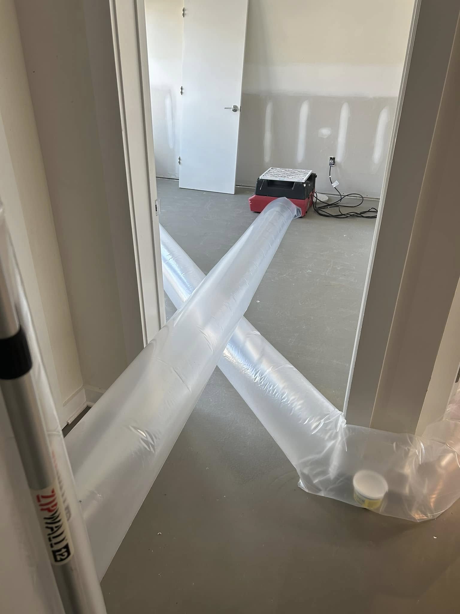 A large plastic ventilation duct is connected to a red machine on the floor of an unfinished room with white walls and an open door.