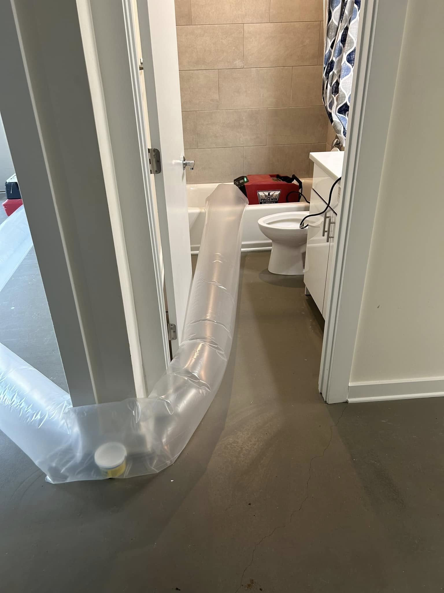 A plastic hose runs from a bathtub, through a bathroom door, and across a concrete floor, connected to equipment near the toilet.
