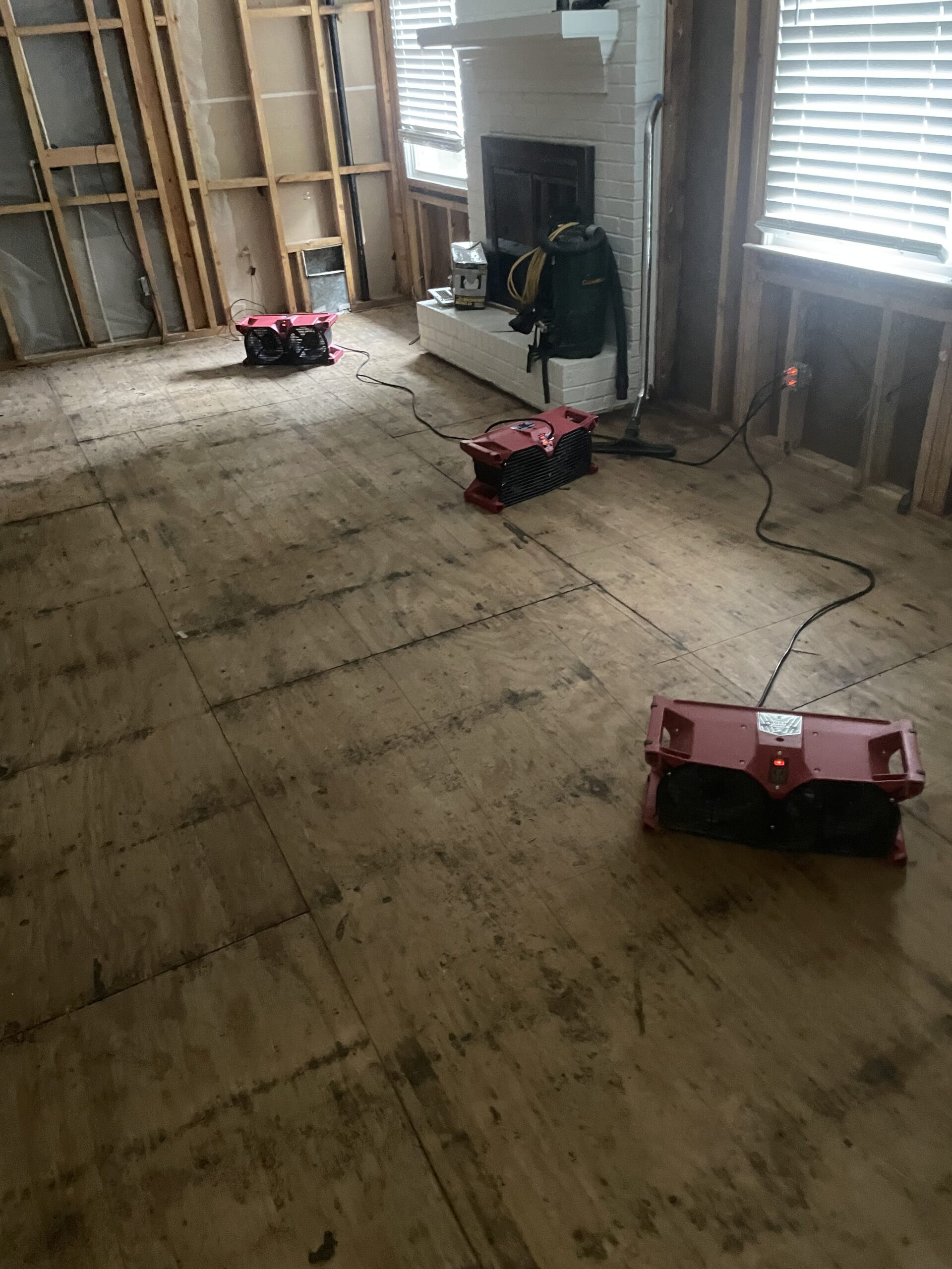 A room with exposed wall studs and plywood flooring, three red drying fans placed on the floor, and a backpack near a white fireplace.