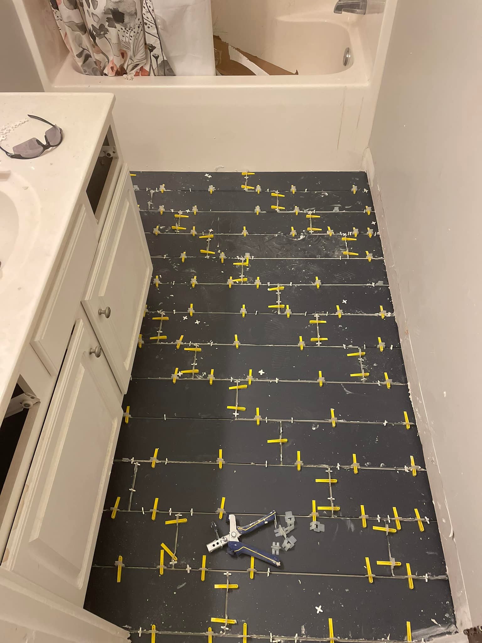 Bathroom floor with newly installed dark tiles, yellow spacers, and tile installation tools; unfinished grout work is visible.