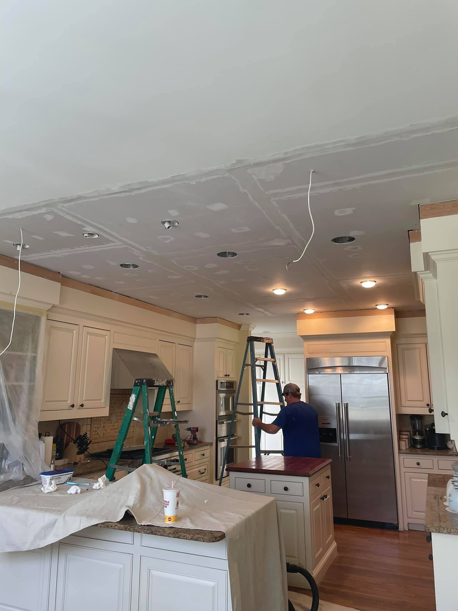 A person stands on a ladder working on a kitchen ceiling with visible drywall seams and exposed wires; tools and drop cloths cover surfaces.
