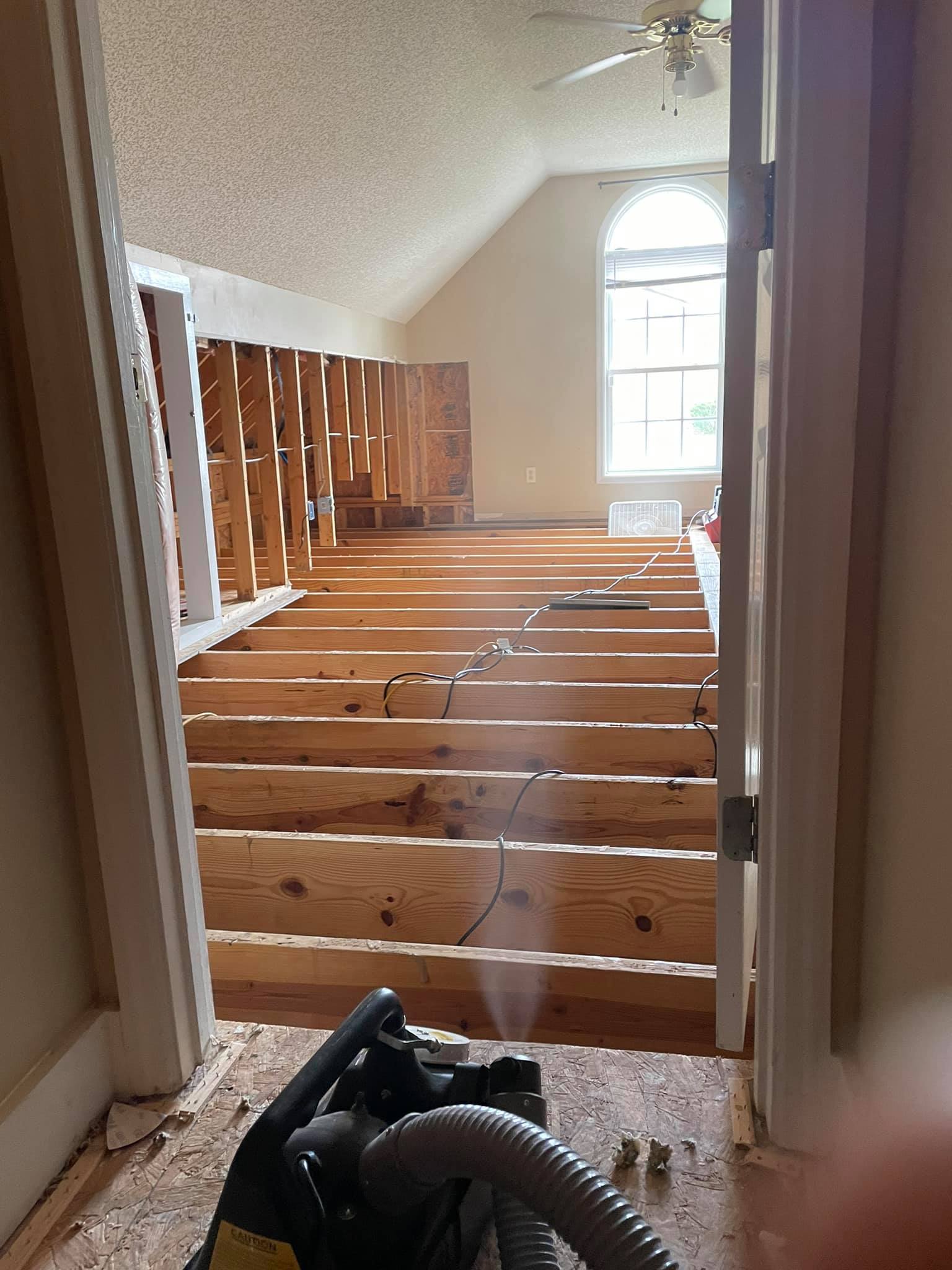Room under renovation with exposed floor joists, visible wiring, and construction equipment; window and ceiling fan in background.