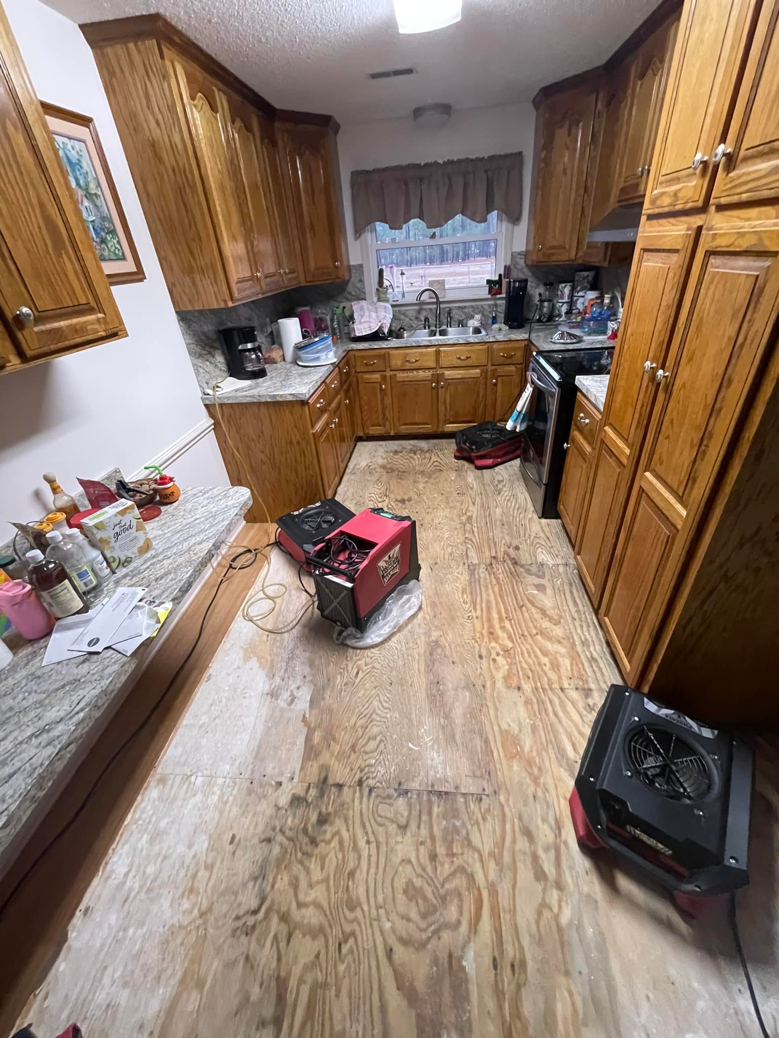 A kitchen with wooden cabinets and no finished flooring; bare plywood subfloor is exposed with drying equipment and tools on the floor.