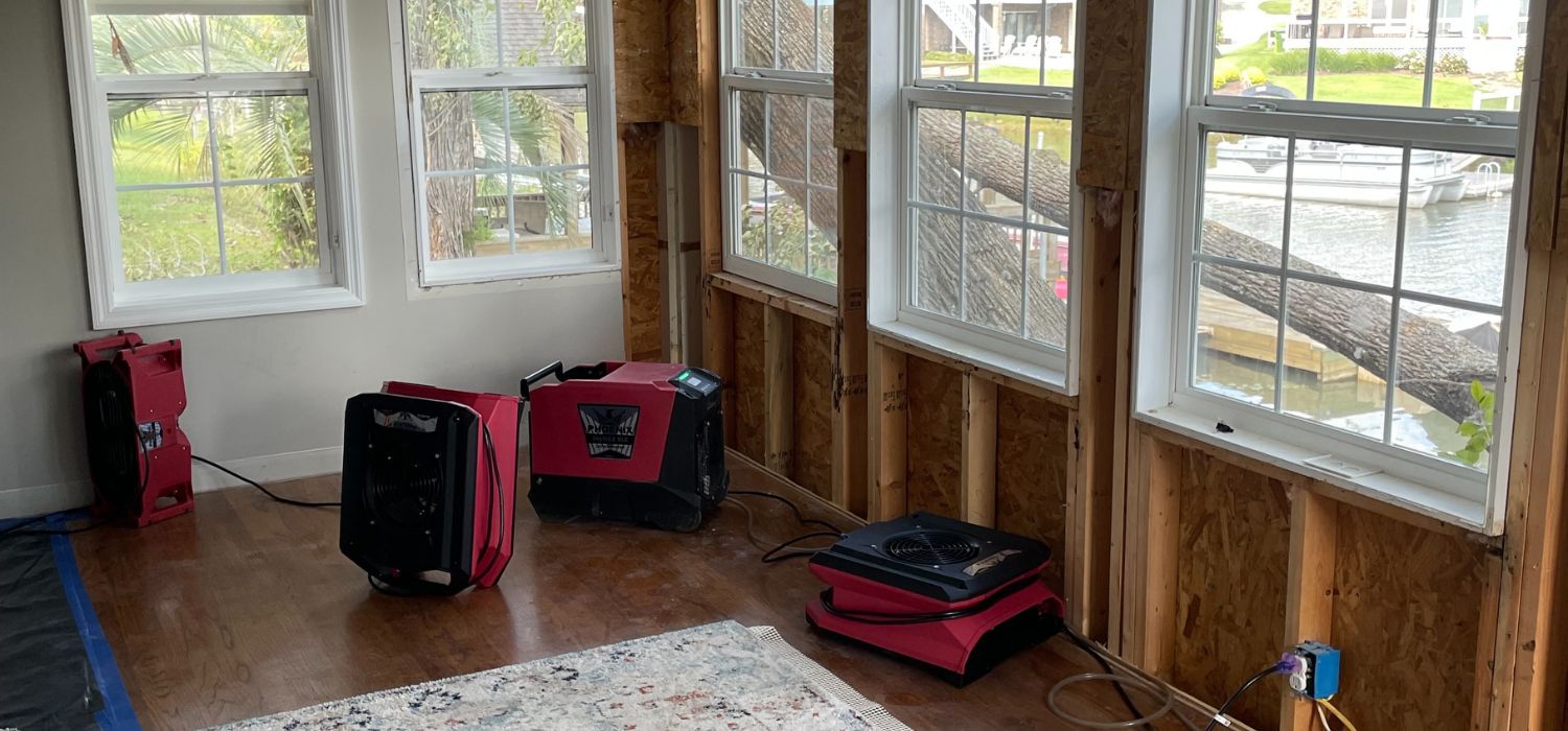 A room with exposed wall studs, three red air movers placed on the floor, and large windows overlooking water outside.
